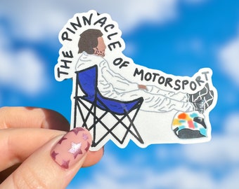 May include: A white sticker with a black line drawing of a person sitting in a blue folding chair. The person is wearing a white jumpsuit and a helmet. The text "THE PINNACLE OF MOTORSPORT" is written in black around the person.