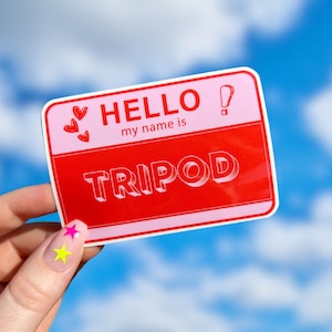 Puede incluir: Una etiqueta de nombre rosa y roja con el texto "HELLO! my name is TRIPOD" y dos corazones rojos. La etiqueta de nombre está sostenida por una mano con dos pegatinas de uñas en forma de estrella amarilla y rosa.