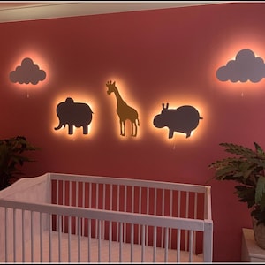 May include: A white crib with a pink wall behind it. There are four light-up wall decorations: a cloud, an elephant, a giraffe, and a hippopotamus. The giraffe is yellow and the other decorations are white.