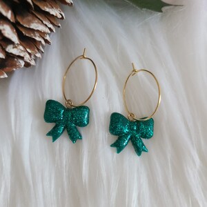 May include: Gold hoop earrings featuring emerald green glitter bow charms. The earrings are presented on a white, fluffy surface, with a pinecone and holly in the background. Festive jewellery.