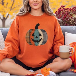 Peut inclure: Un sweat-shirt orange à col rond arborant un motif central de citrouille d'Halloween. Le design de la citrouille est composé de segments verticaux aux motifs écossais et unis dans des tons de marron, bleu et crème avec des yeux noirs.