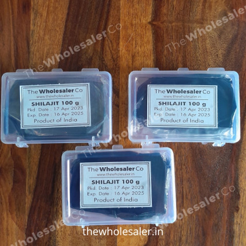 May include: Three clear plastic containers with black lids, each containing a dark brown substance labeled "SHILAJIT 100 g". The label also includes the company name "The Wholesaler Co", website address "www.thewholesaler.in", packaging date, expiration date, and "Product of India".