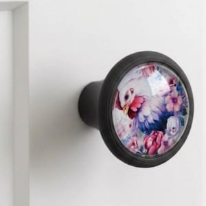 May include: Black cabinet knob with a glass insert showing a colourful rooster and floral design. The knob is round with a black metal base and is mounted on a white surface. The rooster has white, red, and blue feathers.