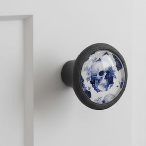 Peut inclure: Un bouton de meuble noir avec une face décorative ronde. La face présente un motif de crâne bleu et blanc, sur un fond floral. Le bouton est monté sur une surface blanche.