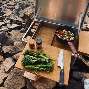 May include: A stainless steel portable kitchen with a wooden work surface. The kitchen is open and shows a variety of cooking supplies, including a pan of vegetables, a knife, and a jar of spices. The kitchen is set up on a rocky beach by a body of water.