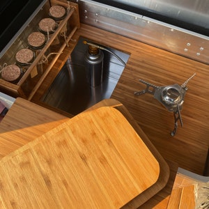 May include: A wooden chopping board with rounded edges sits on top of a wooden surface. A small camping stove is visible in the background.