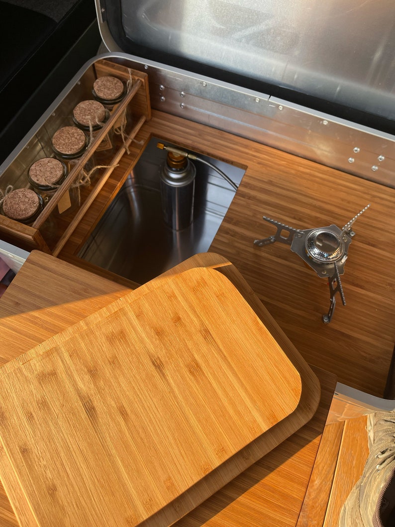 May include: A wooden chopping board with rounded edges sits on top of a wooden surface. A small camping stove is visible in the background.