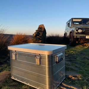 May include: A silver aluminium storage box with a hinged lid and two latches. The box is sitting on a grassy surface with rocks and a green Land Rover in the background.