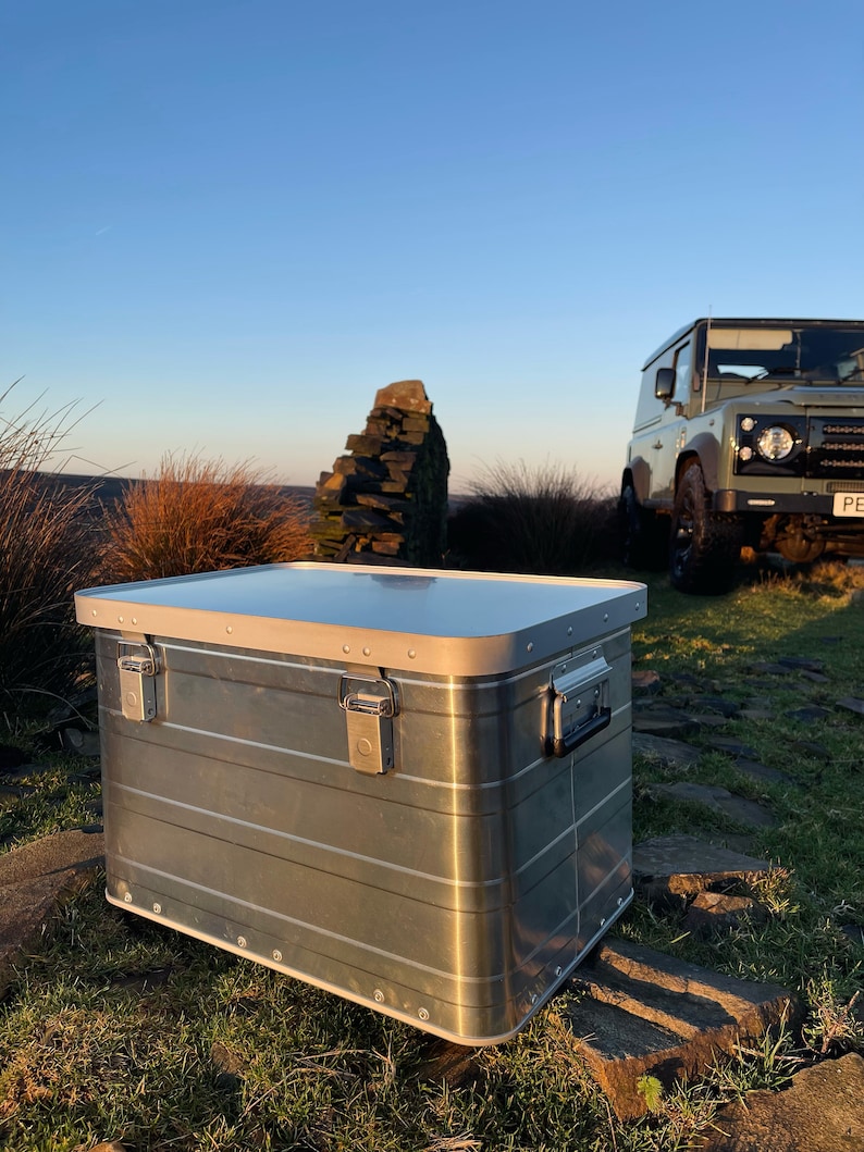 May include: A silver aluminium storage box with a hinged lid and two latches. The box is sitting on a grassy surface with rocks and a green Land Rover in the background.