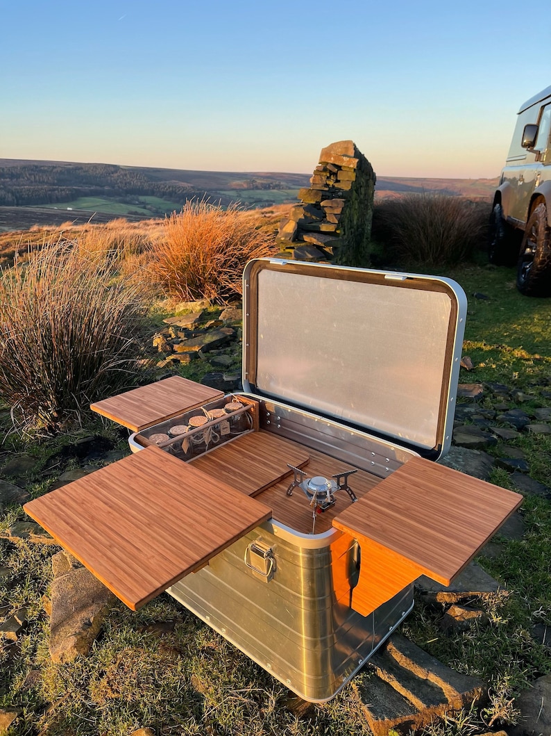 May include: A silver metal camping box with a wooden top and sides that fold out to create a table. The box is open and has a small camping stove inside. The box is sitting on a grassy hill with a stone wall and a view of a valley in the background.
