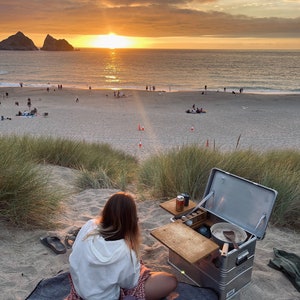 May include: A person sits on a blanket in front of a portable kitchen set up on a sandy beach. The kitchen has a wooden countertop, a silver metal box, and a variety of cooking supplies. The person is wearing a white hoodie and a patterned skirt. The beach is at sunset, with a bright orange sky and a calm sea in the background.