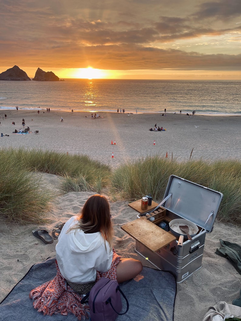 May include: A person sits on a blanket in front of a portable kitchen set up on a sandy beach. The kitchen has a wooden countertop, a silver metal box, and a variety of cooking supplies. The person is wearing a white hoodie and a patterned skirt. The beach is at sunset, with a bright orange sky and a calm sea in the background.