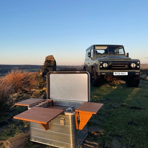 May include: A silver, rectangular, portable kitchen with two wooden fold-out tables. The kitchen is open and has a gas stove and a stainless steel sink. The kitchen is sitting on a rocky surface in front of a green Land Rover Defender with the words "Wild Box" on the back.