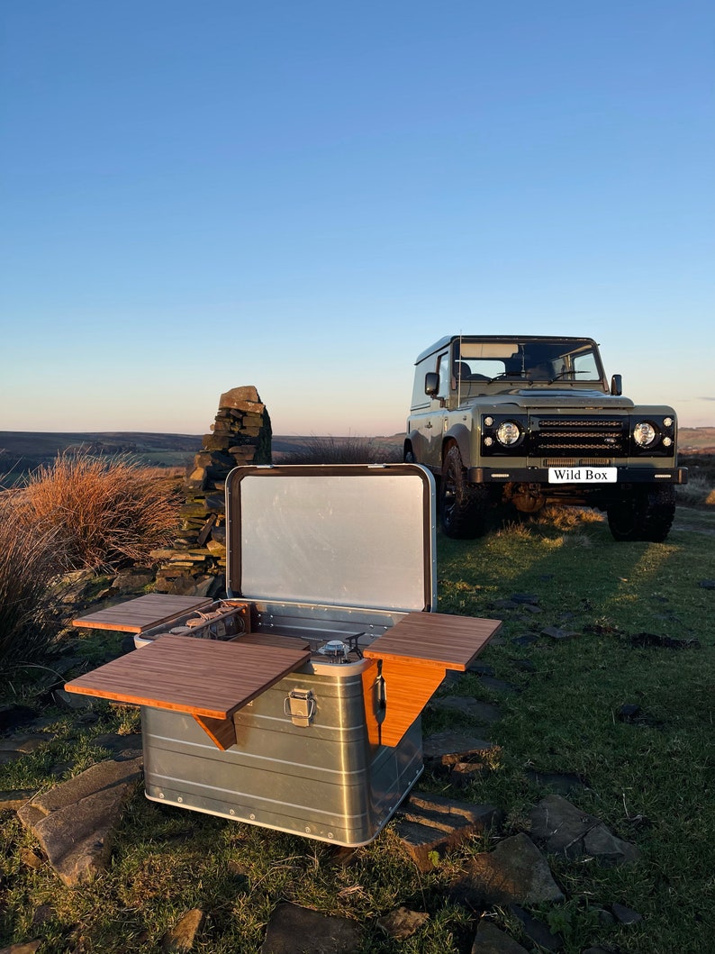 May include: A silver, rectangular, portable kitchen with two wooden fold-out tables. The kitchen is open and has a gas stove and a stainless steel sink. The kitchen is sitting on a rocky surface in front of a green Land Rover Defender with the words "Wild Box" on the back.