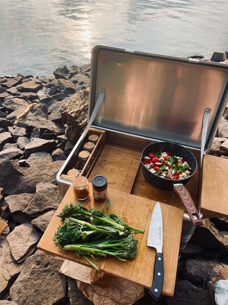 May include: A stainless steel portable kitchen with a wooden work surface. The kitchen is open and shows a variety of cooking supplies, including a pan of vegetables, a knife, and a jar of spices. The kitchen is set up on a rocky beach by a body of water.