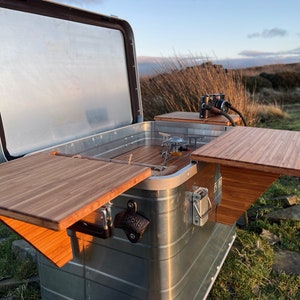 May include: A silver metal storage box with a wooden top and two fold-out wooden side tables. The box is open and the inside is visible. The box is sitting on a grassy surface.