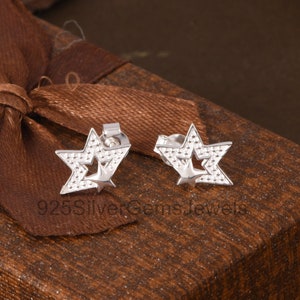 Puede incluir: Un par de pendientes de plata con forma de estrella. Cada pendiente presenta una estrella más grande con una estrella más pequeña recortada en el centro. Los pendientes se muestran sobre una superficie marrón con una cinta.