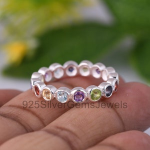 Op de afbeelding: Een zilveren ring met een band van gekleurde edelstenen. De edelstenen zijn in een cirkel gerangschikt en omvatten rood, oranje, geel, groen, blauw en paars.