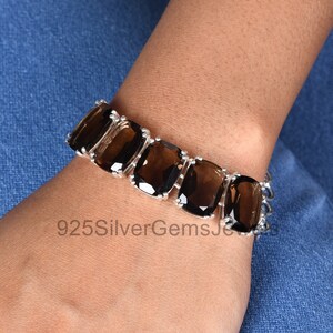 May include: A silver bracelet featuring five large, rectangular, faceted dark brown smoky quartz gemstones set in polished silver prongs. The bracelet is secured with a silver chain link clasp, and the text 925SilverGemsJewels is visible.
