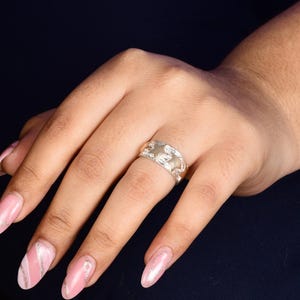 Può includere: Un anello d'argento con un designo testurizzato. L'anello è indossato su un dito con smalto rosa.