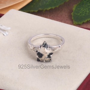 Peut inclure: Une bague en argent avec une pierre précieuse en jaspe dalmatien en forme d'étoile noire et blanche. La bague est posée sur un fond blanc.