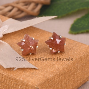 May include: A pair of star-shaped stud earrings featuring faceted sunstone gems set in sterling silver prongs. The earrings are displayed on a textured tan surface with a light ribbon. The text 925SilverGemsJewels is visible in the image.