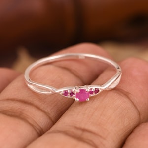 May include: A delicate silver ring featuring a central round-cut pink gemstone flanked by two smaller pink stones on each side. The band has a slender, polished metallic finish with a split design that tapers toward the setting of the stones.