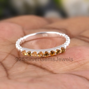 Puede incluir: Un anillo de banda de plata con una fila de pequeñas piedras preciosas redondas de color marrón claro. El anillo está en el dedo de una persona.