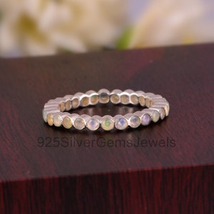 以下が含まれることがあります： 小さな丸い虹色のオパールが並んだシルバーのバンドリング。リングは暗い木製の表面の上に置かれています。