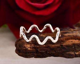 May include: A silver-colored ring with a wavy design. The ring is set against a wooden surface, with a blurred red rose in the background. The ring's design resembles a series of connected waves.