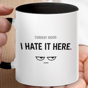 Op de afbeelding: Witte keramische mok met een zwarte binnenkant en handvat. De mok heeft de tekst "CURRENT MOOD: I HATE IT HERE." in het zwart, samen met een fronsende gezichtsafbeelding. De mok wordt door een persoon vastgehouden.