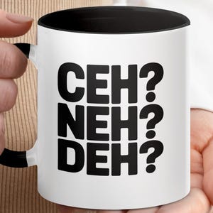 Puede incluir: Taza de cerámica blanca con interior y asa negros. La taza presenta el texto "CEH? NEH? DEH?" en letras negras en negrita. La taza está sujeta por una persona.