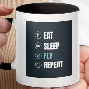 Puede incluir: Taza de cerámica blanca con borde y asa negros. La taza tiene un gráfico en blanco y negro con el texto "Comer, Dormir, Volar, Repetir" e iconos para cada palabra.