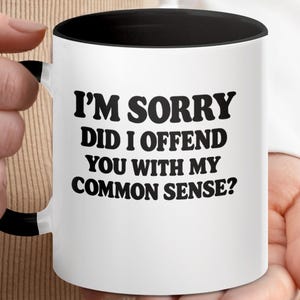 Może przedstawiać: Biały ceramiczny kubek z czarnym wnętrzem i uchwytem. Kubek zawiera tekst "I'M SORRY DID I OFFEND YOU WITH MY COMMON SENSE?" pogrubionymi, czarnymi literami. Kubek jest trzymany przez osobę.