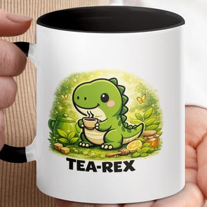 Può includere: Tazza in ceramica bianca con interno e manico neri. La tazza presenta un'illustrazione a cartoni animati di un dinosauro verde che tiene una tazza da tè, con il testo "TEA-REX" sotto. Lo sfondo include una teiera, miele e limone.