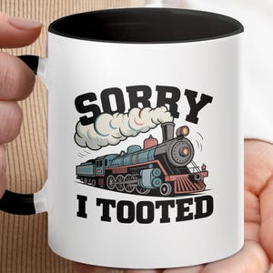 Könnte beinhalten: Weißer Keramikbecher mit schwarzem Innenraum und Griff. Der Becher zeigt eine Cartoon-Illustration einer Dampflokomotive mit dem Text "SORRY I TOOTED" in fetten, schwarzen Buchstaben. Der Zug ist blau, rot und schwarz.