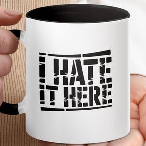 White mug with black handle featuring "I HATE IT HERE" text design.