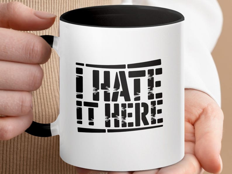 White mug with black handle featuring "I HATE IT HERE" text design.
