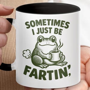 May include: White ceramic mug with a black interior and handle. The mug features a green cartoon frog holding a cup of coffee, with the text "SOMETIMES I JUST BE FARTIN'" in green.