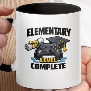 Puede incluir: Taza de cerámica blanca con interior y asa negros. La taza presenta el texto "ELEMENTARY LEVEL COMPLETE" con gráficos de un birrete, un diploma y un mando de videojuegos.