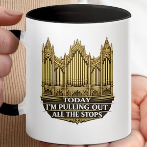 May include: A white ceramic mug with a black interior and handle, featuring a gold-coloured illustration of a pipe organ. Below the organ, the text reads: TODAY I'M PULLING OUT ALL THE STOPS, printed in white capital letters on a dark background.