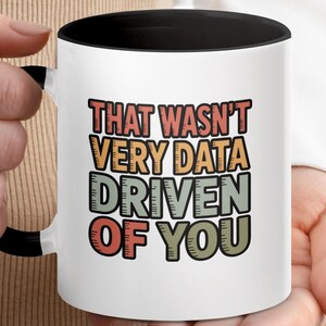 Könnte beinhalten: Weißer Keramikbecher mit schwarzem Innenraum und Henkel. Der Becher zeigt den Text "THAT WASN'T VERY DATA DRIVEN OF YOU" in einer Retro-Schriftart in mehreren Farben. Der Becher wird von einer Person gehalten.