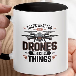 以下が含まれることがあります： 黒い内側とハンドルが付いた白いセラミックマグ。テキスト「THAT'S WHAT I DO、I FLY、DRONES、AND I KNOW THINGS」とドローンのイラストが特徴です。マグカップは人が持っています。