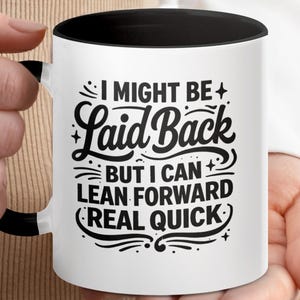 May include: White ceramic mug with a black interior and handle. The mug features the text "I MIGHT BE Laid Back BUT I CAN LEAN FORWARD REAL QUICK" in a black, stylized font. The mug is held by a person.