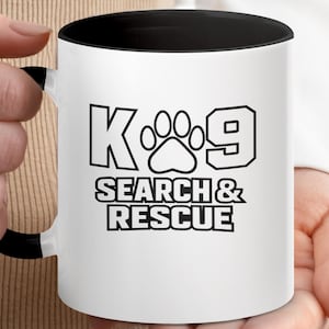 May include: A white ceramic mug with a black interior and a black handle. The front of the mug features a black graphic design with the text K-9 and a paw print icon, followed by the text SEARCH & RESCUE in bold, block capital letters.