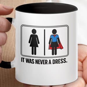 May include: White ceramic mug with a black interior and handle. The mug features a graphic of two stick figures, one in a dress and the other in a superhero costume, with the text "IT WAS NEVER A DRESS."