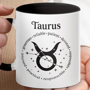 Peut inclure: Mug en céramique blanche avec intérieur et anse noirs, avec le mot "Taurus" et un symbole de taureau noir avec des détails de constellation. Le design comprend des mots comme "fiable" et "dévoué".