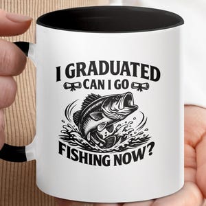 Può includere: Una tazza in ceramica bianca con interno e manico neri, caratterizzata da una grafica nera di un pesce che salta e due diplomi di laurea. Il testo stampato sulla tazza recita: I GRADUATED CAN I GO FISHING NOW? Il design è centrato sul davanti.