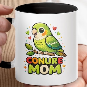 Könnte beinhalten: Weißer Keramikbecher mit schwarzem Innenraum und Henkel. Der Becher zeigt eine Cartoon-Illustration eines grünen Conure-Papageis, der auf einem Ast sitzt, mit den Worten "CONURE MOM" in bunten Buchstaben. Herzen und Punkte umgeben den Vogel.