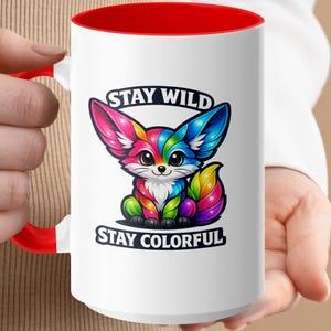 White mug with red rim featuring a colorful cartoon fox and text "STAY WILD STAY COLORFUL".